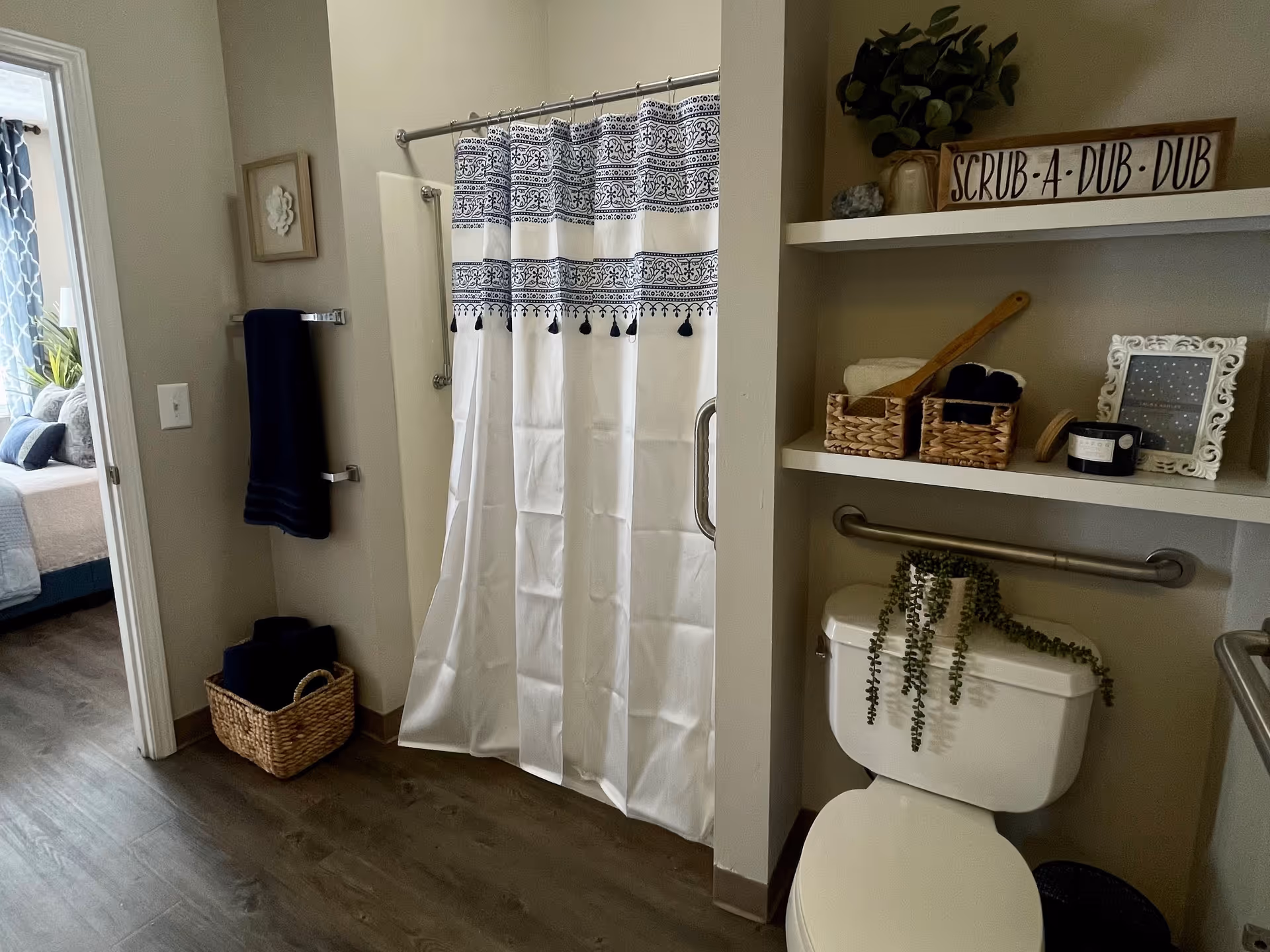 Bathroom with a shower curtain, toilet topped with a trailing plant, wall shelves holding baskets and decor, and grab bars.