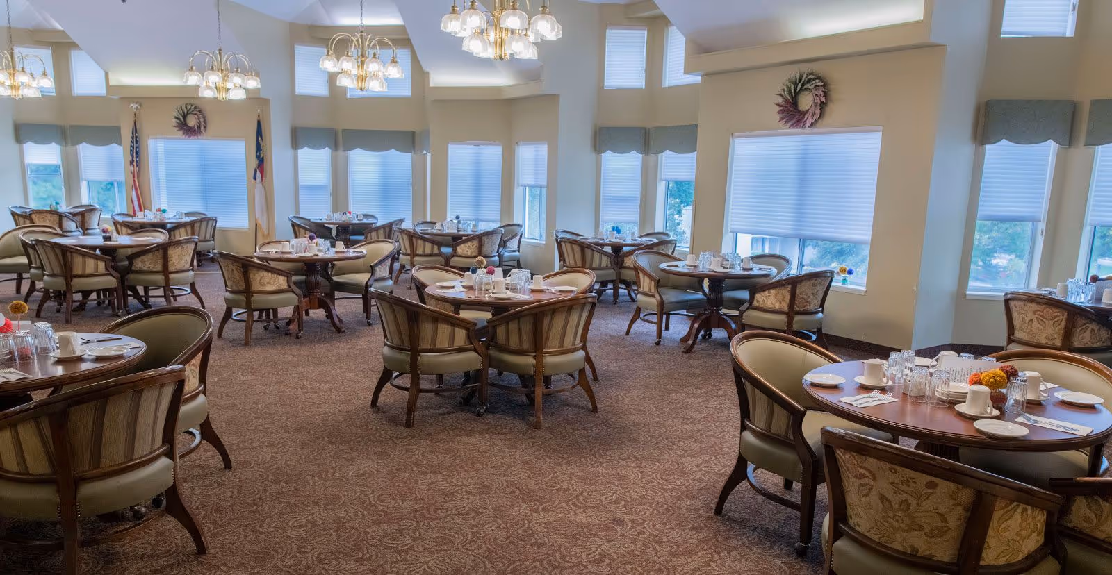 Spacious dining room with multiple round tables set for meals and upholstered chairs beneath chandeliers.