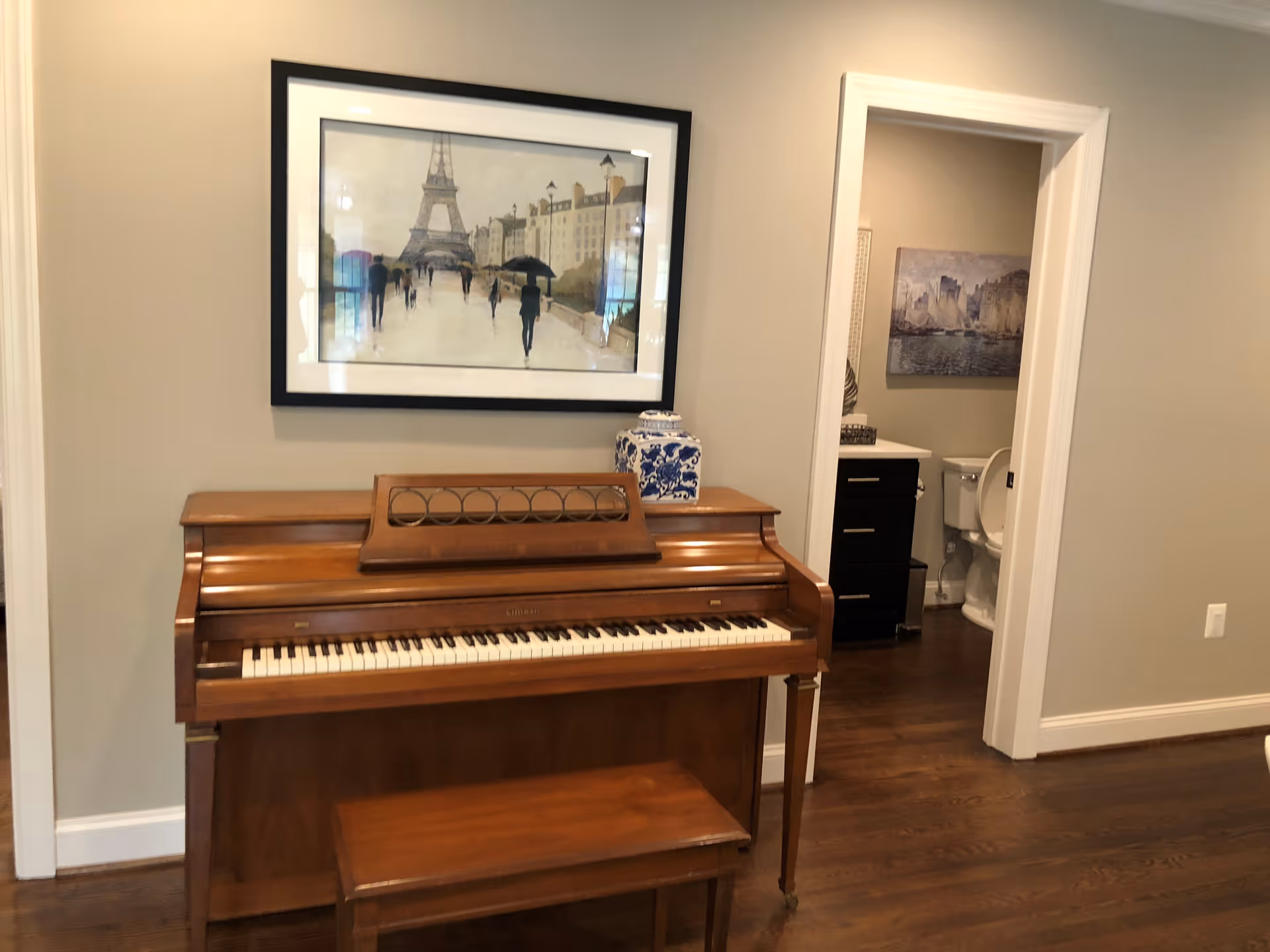 A wooden piano with a matching bench is placed against a beige wall. Above the piano hangs a framed painting depicting people walking near the Eiffel Tower. To the right, there is an open doorway leading to a bathroom with a toilet, a dark-colored vanity with a white countertop, and a painting of boats on the wall. The floor is made of dark wood.