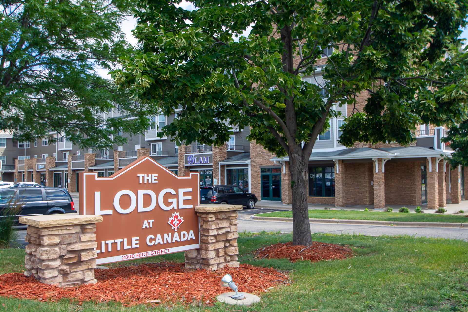 Outdoor view of The Lodge at Little Canada sign with a tree and landscaped area in the foreground, and a multi-story building with storefronts and parked cars in the background.