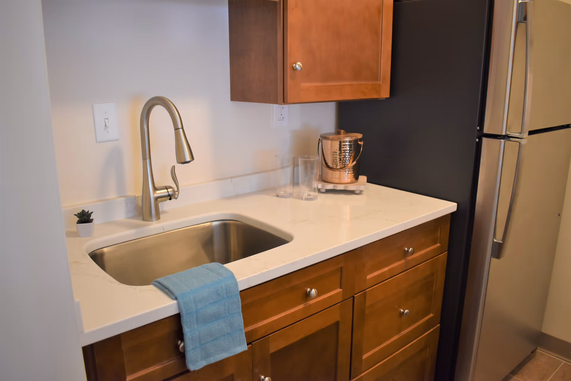 A kitchen countertop with a stainless steel sink and a modern faucet. A blue towel is draped over the edge of the sink. There are wooden cabinets below and above the countertop. On the countertop, there is a small potted plant, two clear glasses, and a copper ice bucket. A stainless steel refrigerator is visible on the right side.