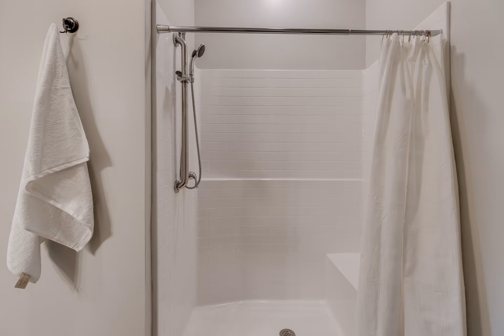 A clean, white shower area with a built-in bench, a handheld showerhead attached to a vertical grab bar, a white shower curtain partially drawn to the side, and a white towel hanging on a hook on the wall.