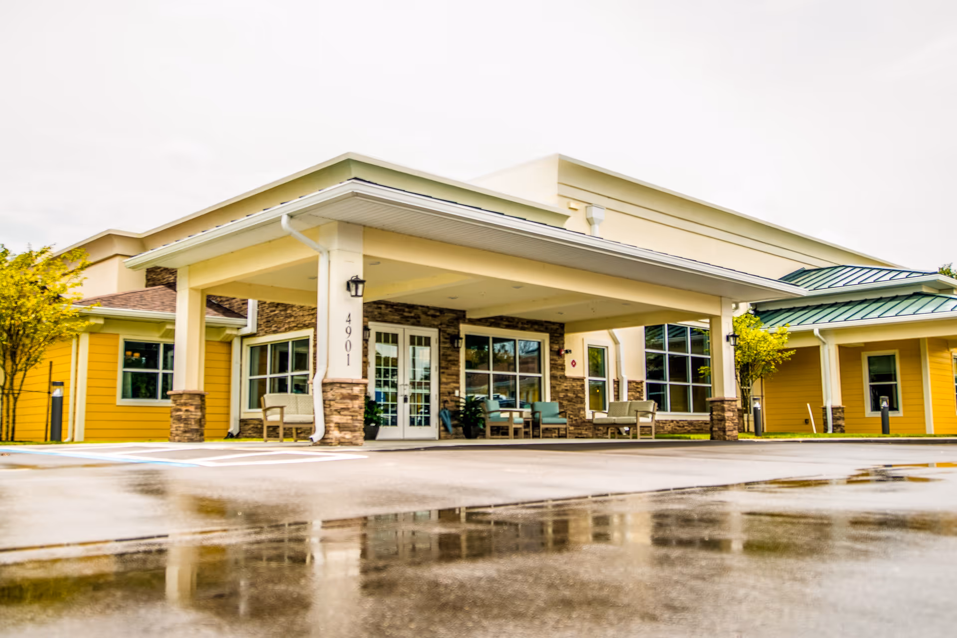 Front exterior view of CERTUS Premier Memory Care Living facility showing a covered entrance with seating areas, yellow walls, stone accents, and large windows. The ground is wet, reflecting the building and surroundings.