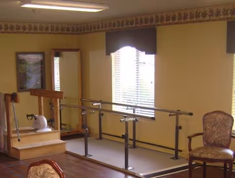 A therapy or rehabilitation room with parallel bars for walking exercises, a small set of steps with a handrail, a large mirror on the wall, and a window with blinds and a valance. There are upholstered chairs with wooden frames in the room, and the walls are painted yellow with a decorative border near the ceiling.