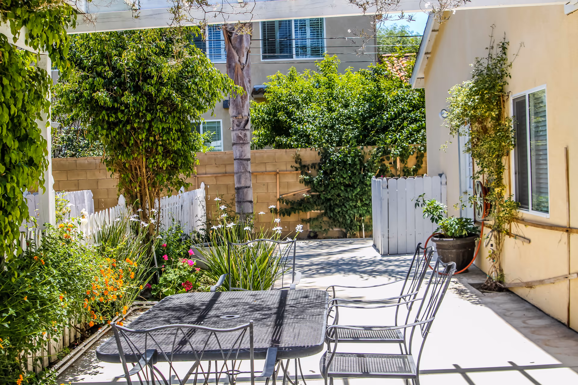 Sunlit courtyard patio with a metal table and chairs, potted plants and flowering shrubs next to a beige building.
