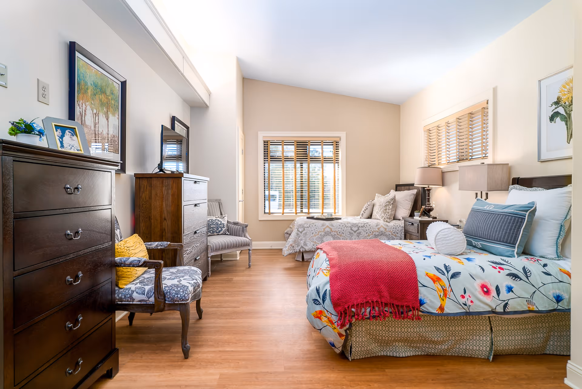 A bright and spacious bedroom with two beds, one with floral bedding and a red throw blanket, and the other with neutral patterned bedding. The room features wooden flooring, two wooden dressers, a TV on one dresser, two upholstered chairs with patterned cushions, and windows with wooden blinds. The walls are decorated with framed artwork and a small plant on one dresser.