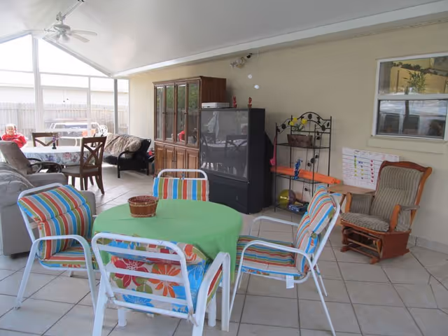 Covered patio area with a round table covered by a green tablecloth surrounded by four colorful striped chairs. In the background, there is a wooden cabinet, an older style television, a metal shelf with various items, a rocking chair, and a window. There is also a dining table with chairs and a person sitting in the far left corner. The floor is tiled and the space is enclosed with a fence visible outside.