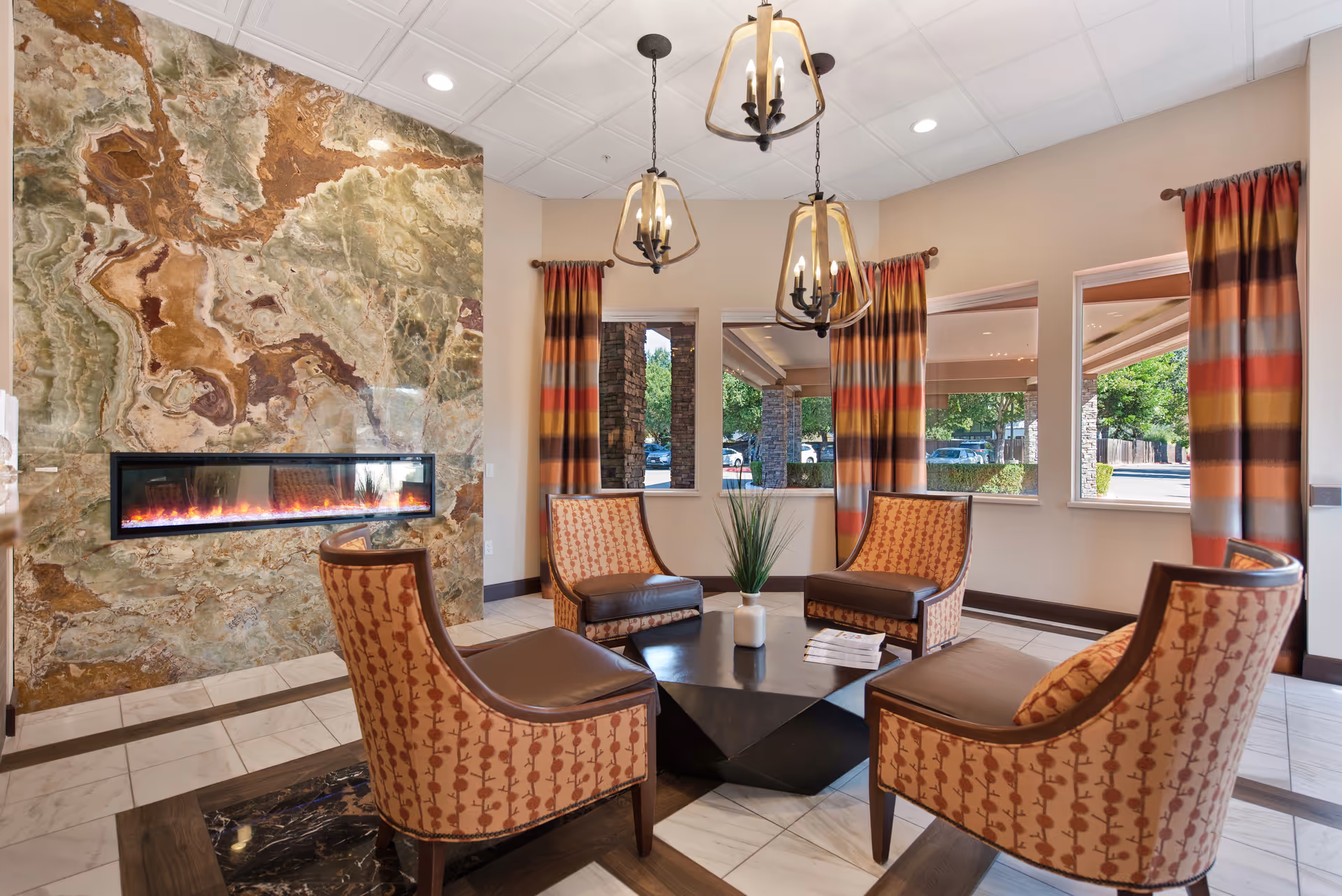 A cozy sitting area in a senior living facility featuring four patterned armchairs arranged around a black geometric coffee table with a small plant and some papers on it. The room has a large decorative stone wall with a modern horizontal electric fireplace. Three pendant lights hang from the ceiling, and windows with colorful striped curtains provide a view of the outside parking area and greenery.