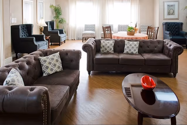 A well-lit living room with two brown tufted sofas adorned with patterned cushions, a dark wooden oval coffee table with a red decorative bowl, and additional seating including two dark armchairs and two patterned chairs near a round table with a pink tablecloth and a basket centerpiece. Light filters in through large windows with sheer curtains.