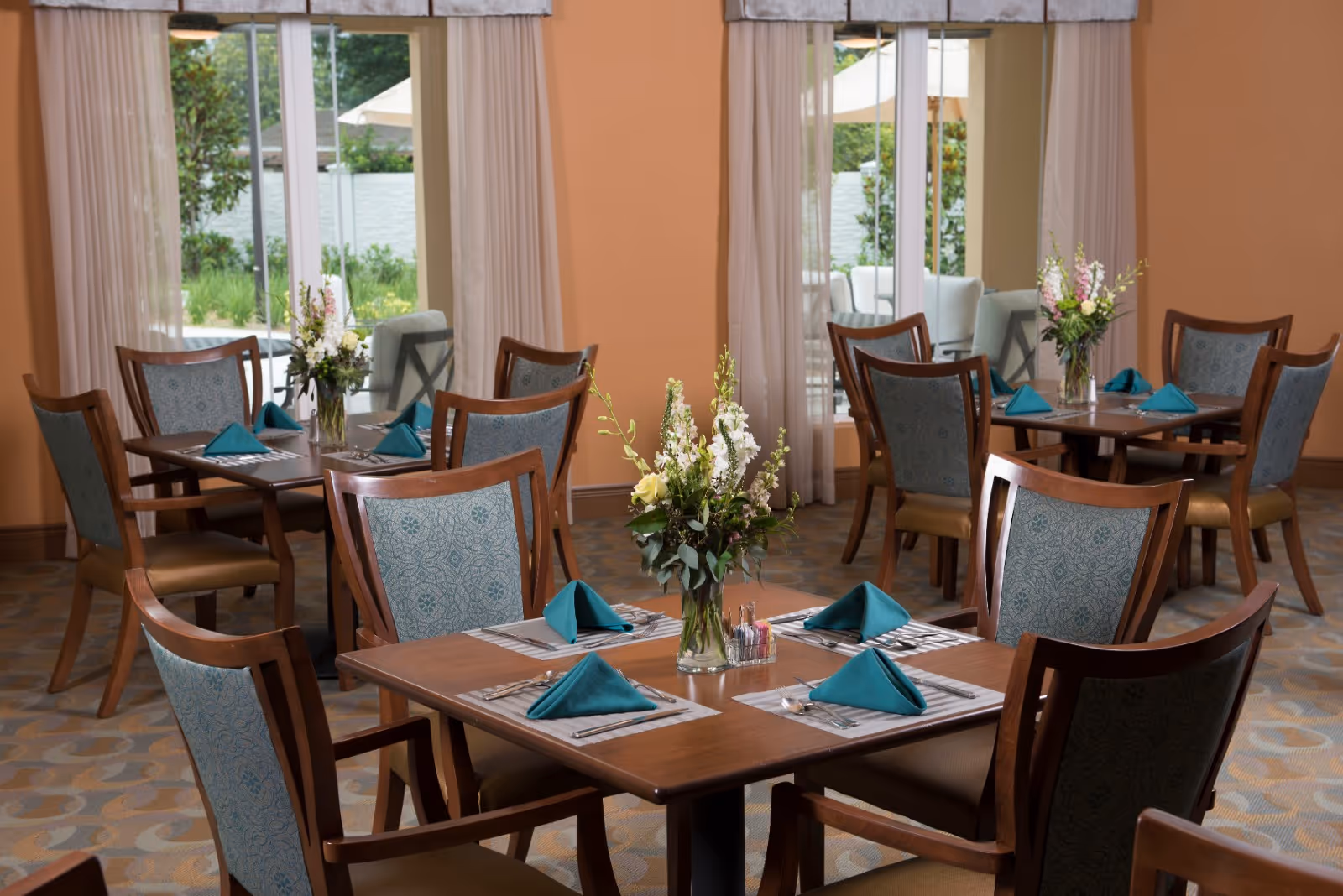 A dining room with several wooden tables and chairs arranged neatly. Each table is set with teal folded napkins, placemats, and silverware. There are vases with fresh flowers on the tables. Large windows with sheer curtains allow natural light to brighten the room and offer a view of an outdoor patio area with greenery.