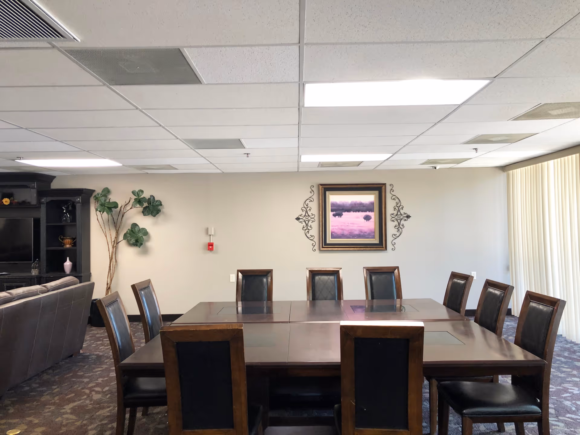 A meeting or dining room with a large rectangular wooden table surrounded by nine chairs with black cushioned seats and backs. The room has a patterned carpet, beige walls, and a ceiling with fluorescent lights. On the wall is a framed painting of a purple landscape with bison, flanked by decorative metal wall art. To the left, there is a dark wooden entertainment unit with shelves and a TV, and a tall plant with large green leaves. Vertical blinds cover the windows on the right side of the room.
