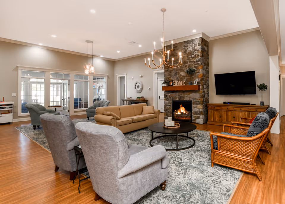 Spacious communal living room with sofas and armchairs arranged around a coffee table, a stone fireplace, wall-mounted TV, and chandelier lighting.