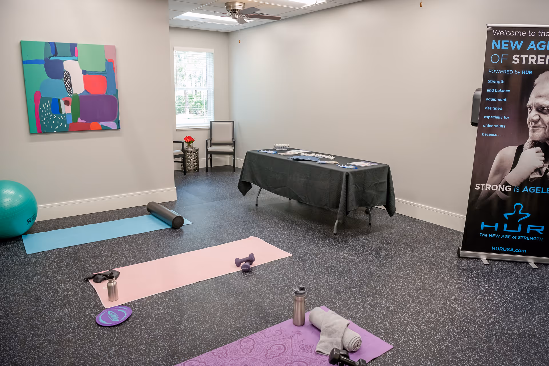 A fitness or exercise room with three yoga mats on the floor, each with different exercise equipment such as dumbbells, a foam roller, a water bottle, and a towel. There is a large green exercise ball in the corner, a colorful abstract painting on the wall, and a table covered with a black cloth holding brochures and water bottles. A standing banner promoting strength equipment for older adults is visible on the right side of the room.