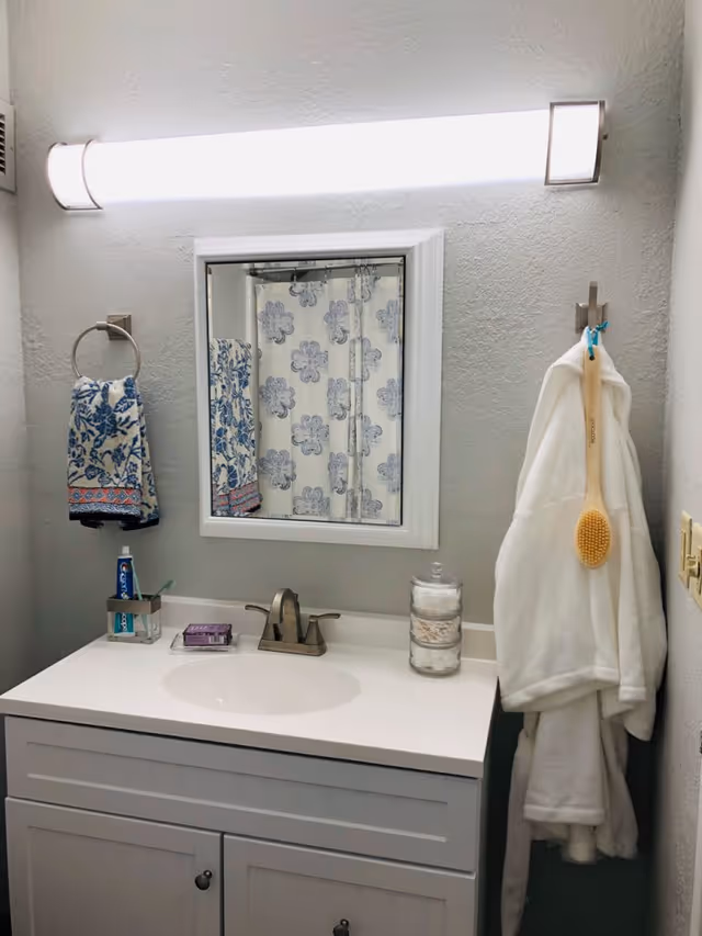 Bathroom vanity with sink and mirror, a patterned towel, a floral shower curtain reflected in the mirror, and a white bathrobe hanging on the wall.
