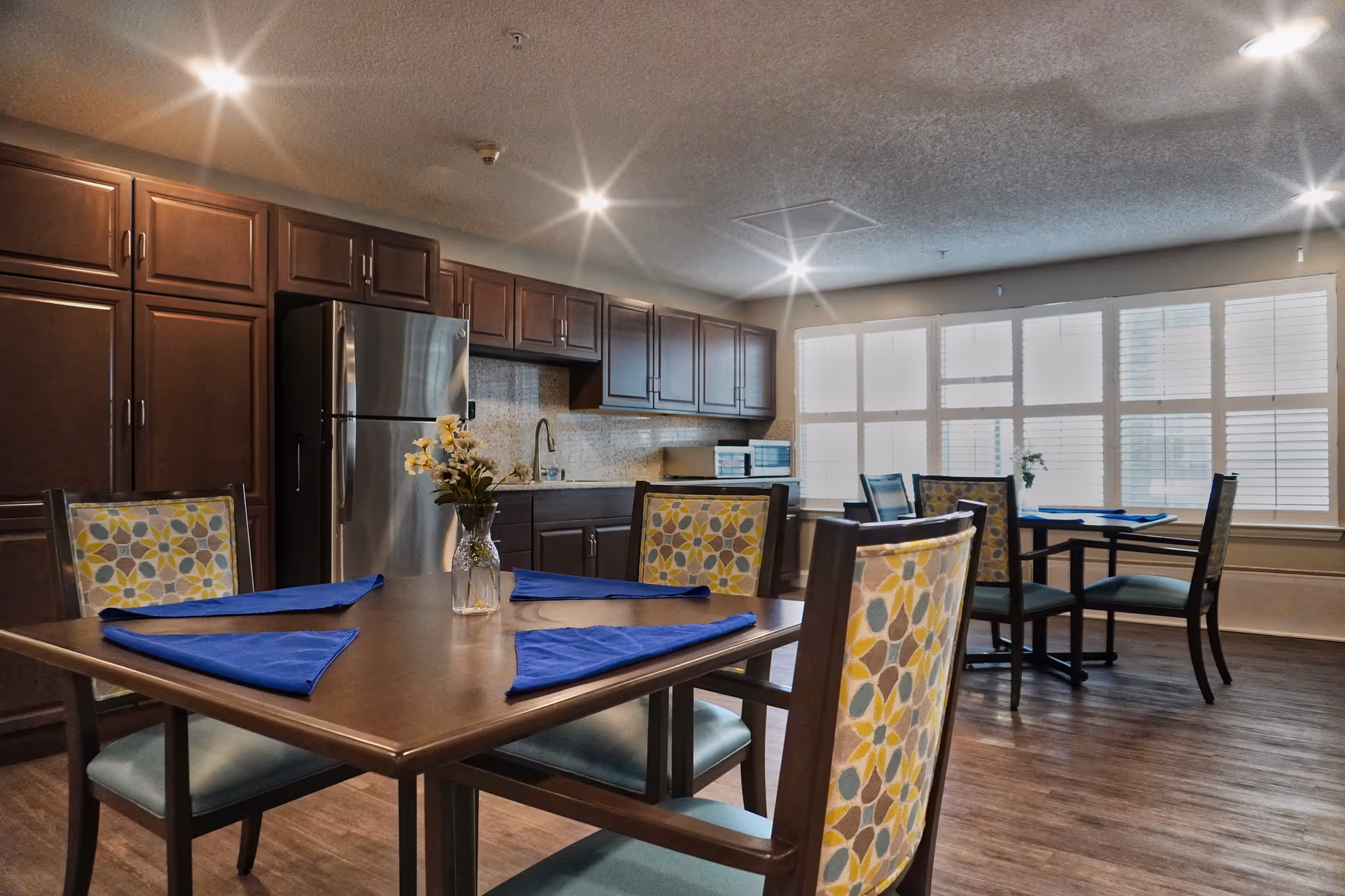 Communal dining area with wooden tables set with blue napkins, patterned chairs, and a kitchenette with dark cabinets and a stainless refrigerator.