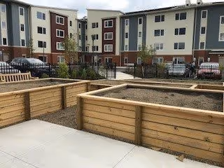 Outdoor area of a senior living facility with raised wooden garden beds, a concrete walkway, and a multi-story residential building in the background.