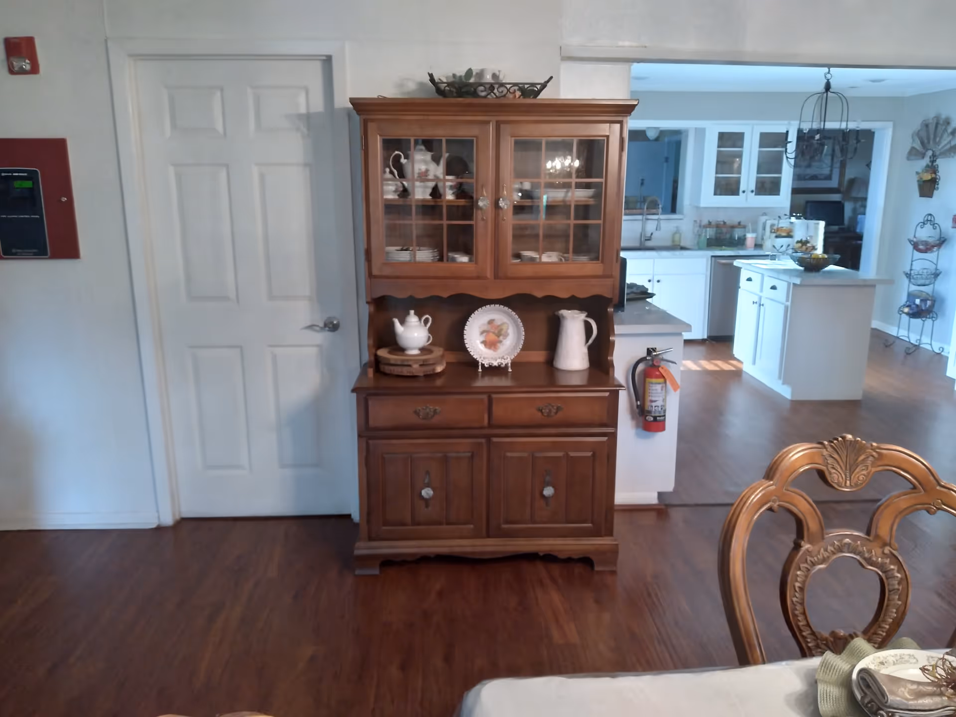 Interior view of a senior living facility showing a wooden china cabinet with dishes and decorative items, a white door on the left, and a kitchen area with white cabinets and an island in the background. Part of a dining table with a decorative chair is visible in the foreground.