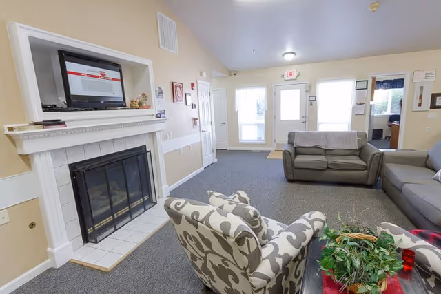 A senior living facility common area with a fireplace on the left wall, a mounted TV above it, and several seating options including two patterned armchairs and two gray sofas. The room has beige walls, carpeted floor, and a door with windows letting in natural light at the far end.