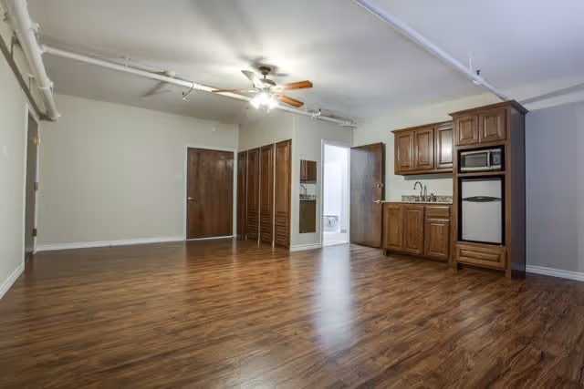 An empty room with wood flooring, a ceiling fan with lights, a small kitchenette with wooden cabinets, a mini fridge, and a microwave. There are two closed wooden doors and a closet with louvered doors. The walls are painted light beige and there is a small bathroom visible through an open door.
