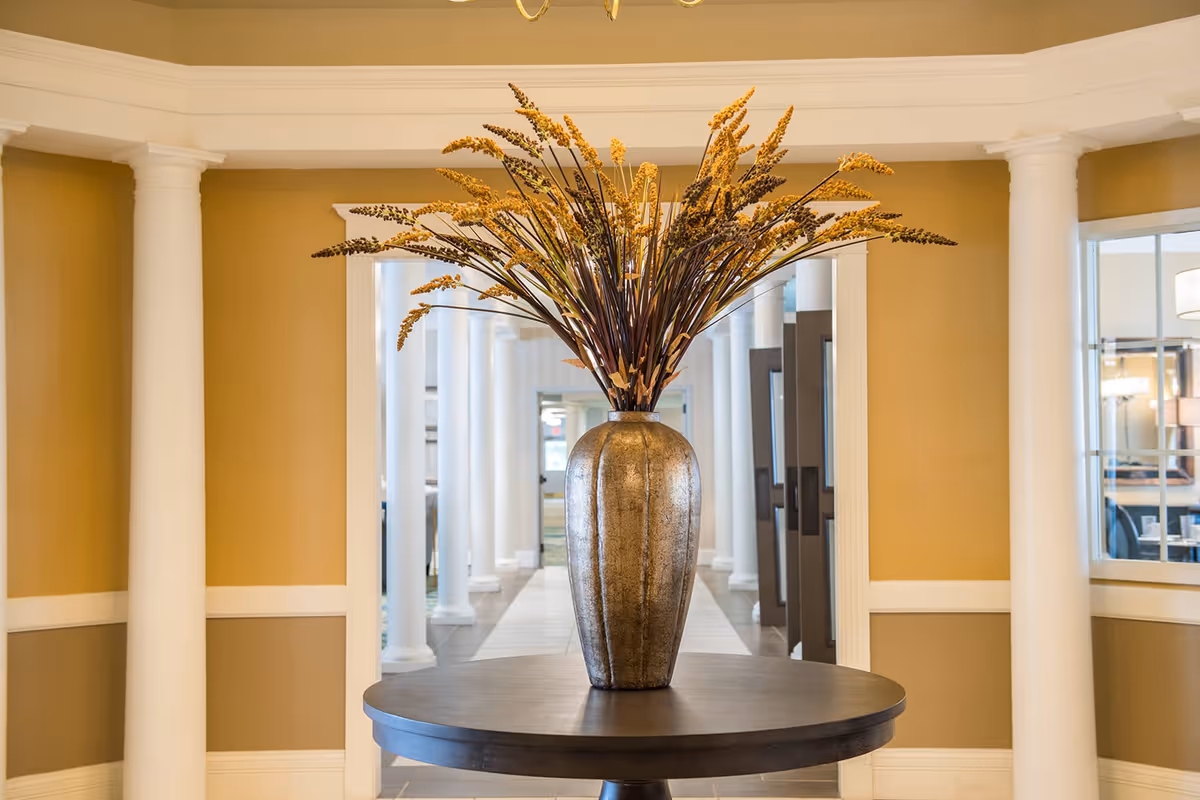 A large decorative vase with tall dried floral arrangements sits on a round wooden table in a hallway with white columns and beige walls, leading to a bright interior space.