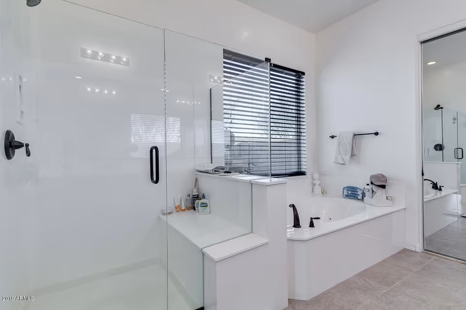 Bright modern bathroom with a glass-enclosed shower on the left, a white bathtub with black fixtures on the right, a window with black blinds above the bathtub, and a large mirror reflecting part of the room.