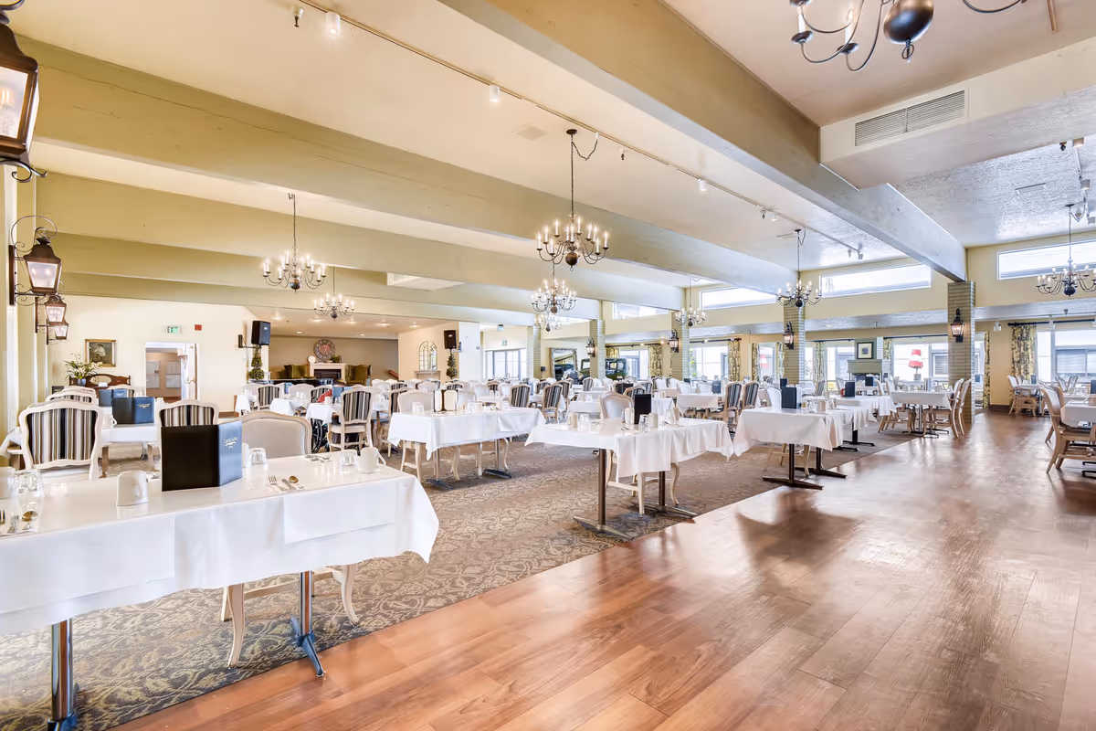 A spacious and well-lit dining room in an assisted living community with multiple tables covered in white tablecloths, set with glasses, cups, and menus. The room features chandeliers hanging from the ceiling, large windows allowing natural light, and a mix of carpeted and wooden flooring.