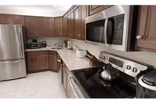 Modern kitchen with stainless steel appliances including a refrigerator, microwave, and electric stove. The kitchen features dark wood cabinets, a light-colored granite countertop, and various small appliances such as a toaster and coffee maker.