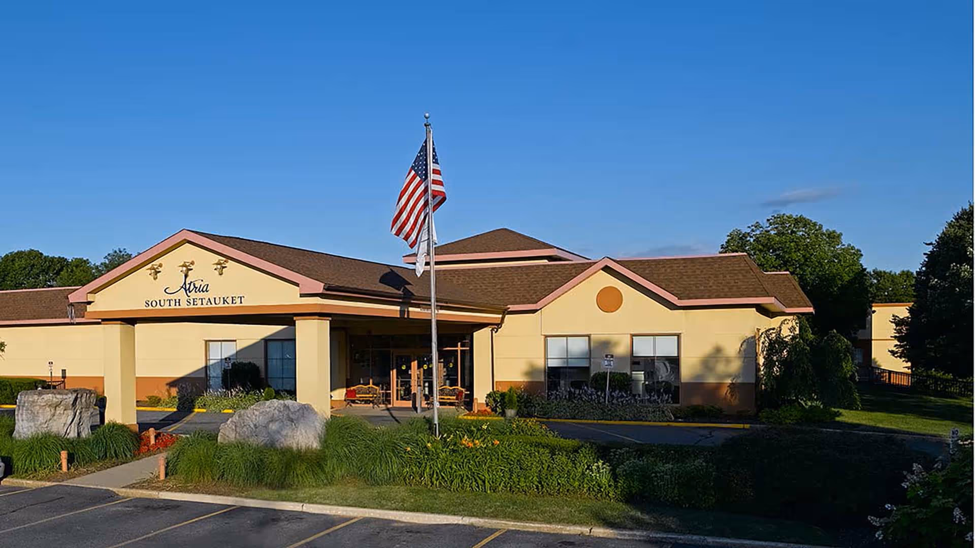 Front entrance of a senior living community building with an American flag, covered porte-cochere, and landscaped rock garden.