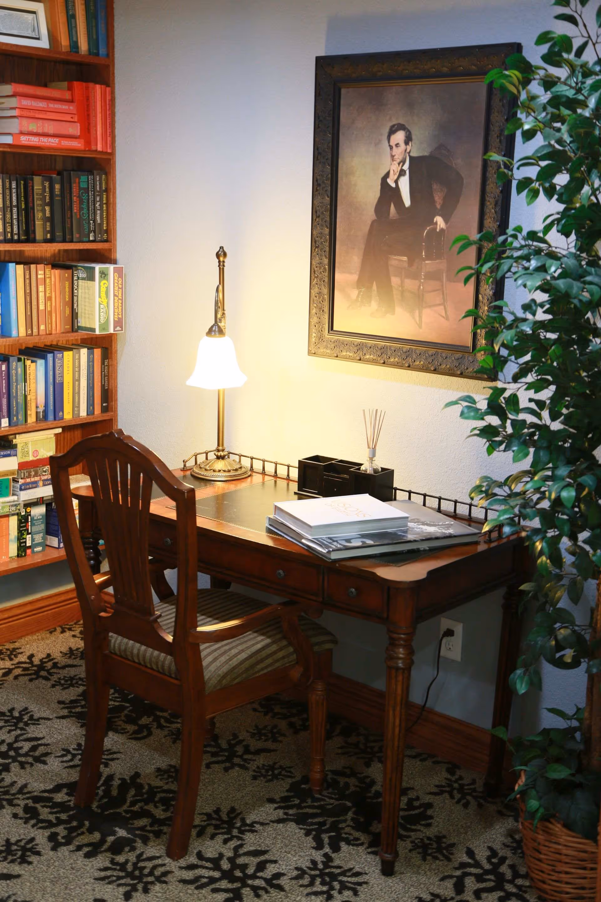 A wooden desk and chair in a cozy reading nook with a lamp, bookshelves, a framed portrait on the wall, and a potted plant.