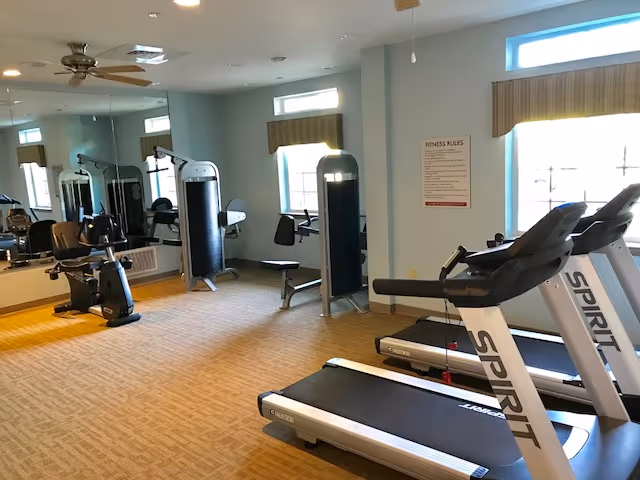 Fitness room with exercise equipment including two Spirit treadmills, a recumbent bike, weight machines, and a bench. The room has beige carpet, light blue walls, large windows with valances, a ceiling fan, and a large mirror on one wall.