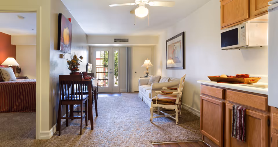 Interior view of a senior living facility apartment at Acacia Springs showing a small kitchen area with wooden cabinets and a microwave, a living room with a sofa, armchair, lamp, and framed artwork on the wall, a dining area with a table and chairs, and a bedroom partially visible through an open doorway. French doors at the far end lead outside, allowing natural light into the space.