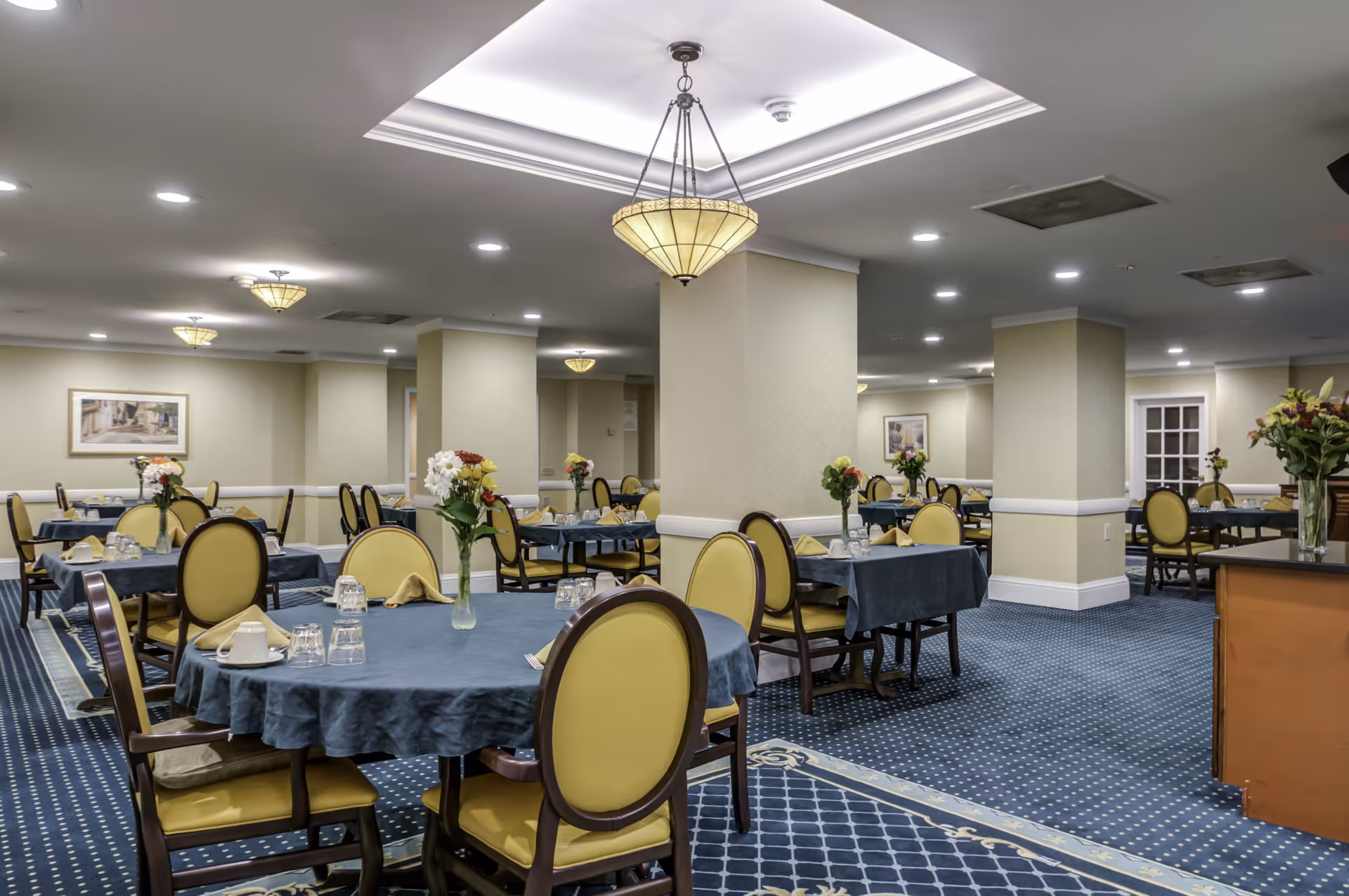 A spacious dining room with multiple round and rectangular tables covered with blue tablecloths. Each table is set with glasses, napkins, and a vase of flowers. The room has yellow cushioned chairs, blue patterned carpet, beige walls, and ceiling lights including a central chandelier.