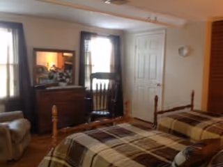 A bedroom with two single beds covered in plaid bedspreads, a wooden dresser with a mirror, a wooden chair, and two windows with curtains allowing natural light into the room.