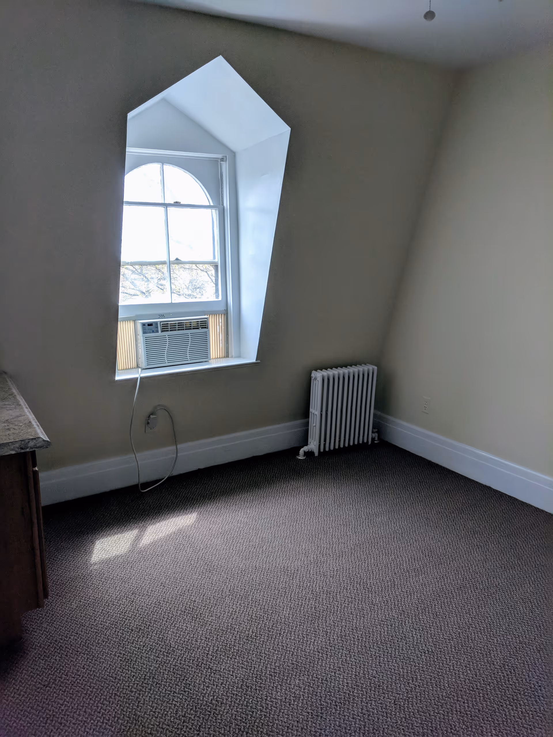 Empty carpeted room with a dormer arched window fitted with a window air conditioner and a radiator against beige walls.