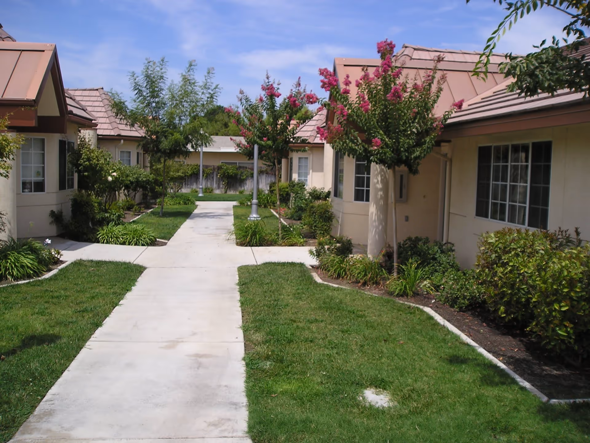 A paved walkway flanked by green lawns and landscaped bushes and trees, leading between single-story buildings with beige walls and brown roofs under a blue sky.