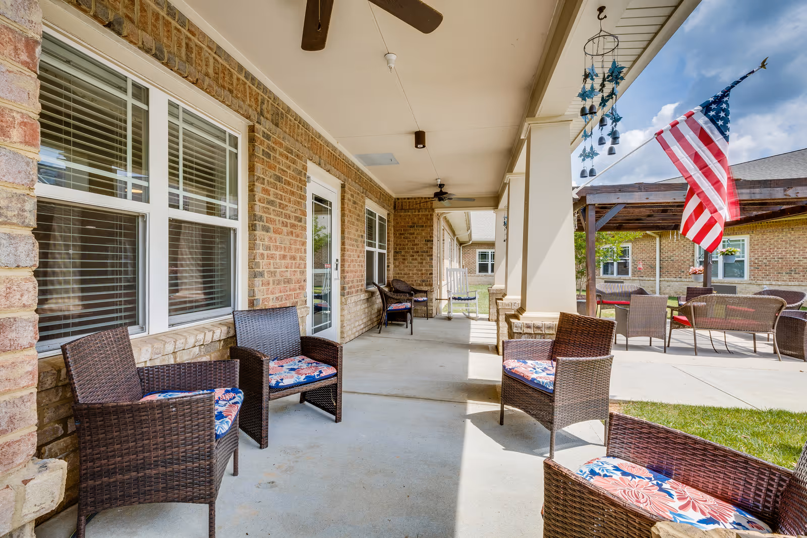 Covered outdoor patio area at Chatham Ridge Assisted Living with wicker chairs featuring floral cushions, ceiling fans, a hanging wind chime, and an American flag. The patio overlooks a grassy area and additional seating under a wooden pergola.
