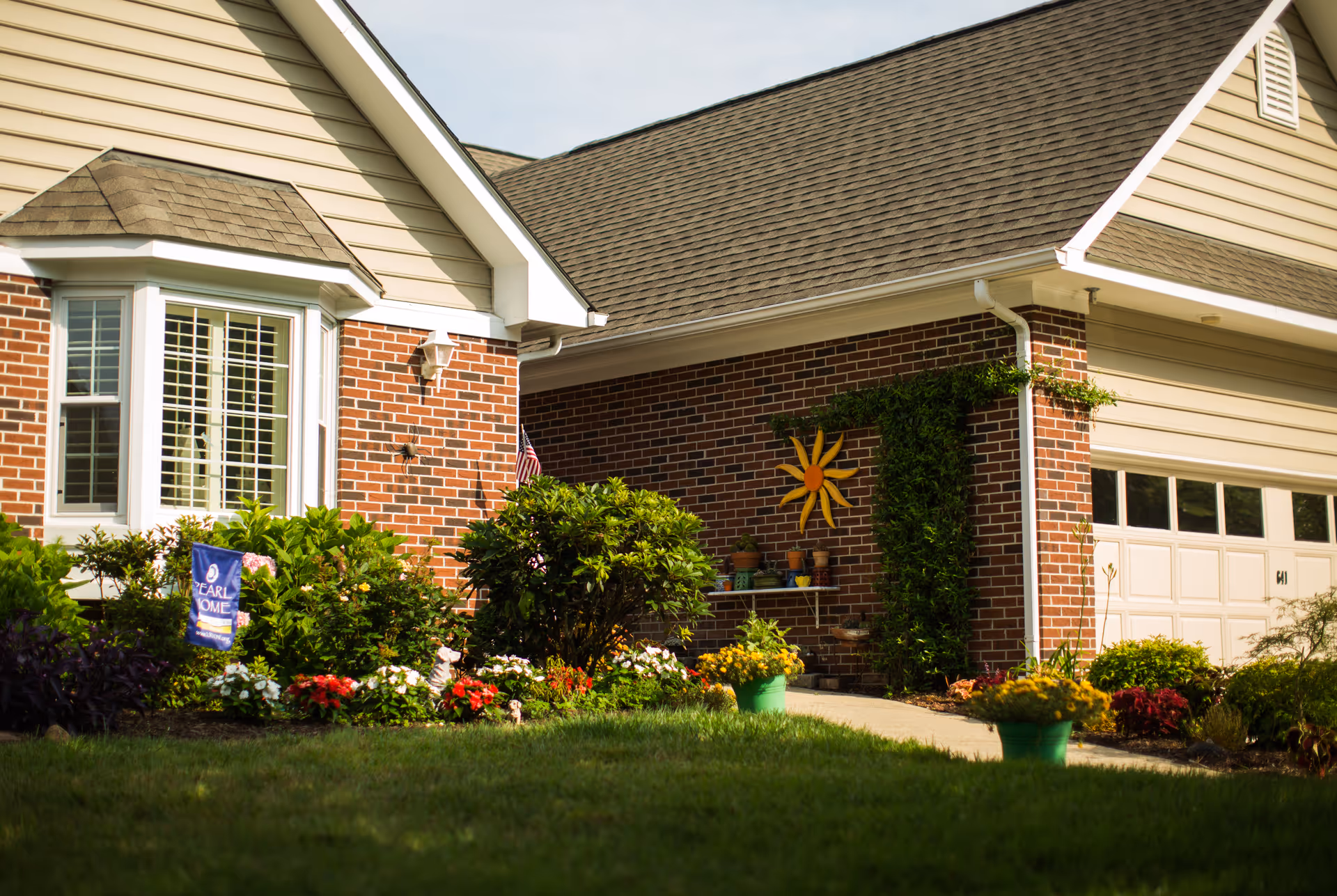 Front exterior of a brick suburban home with a garage, bay window, flower beds and a decorative sun on the wall.
