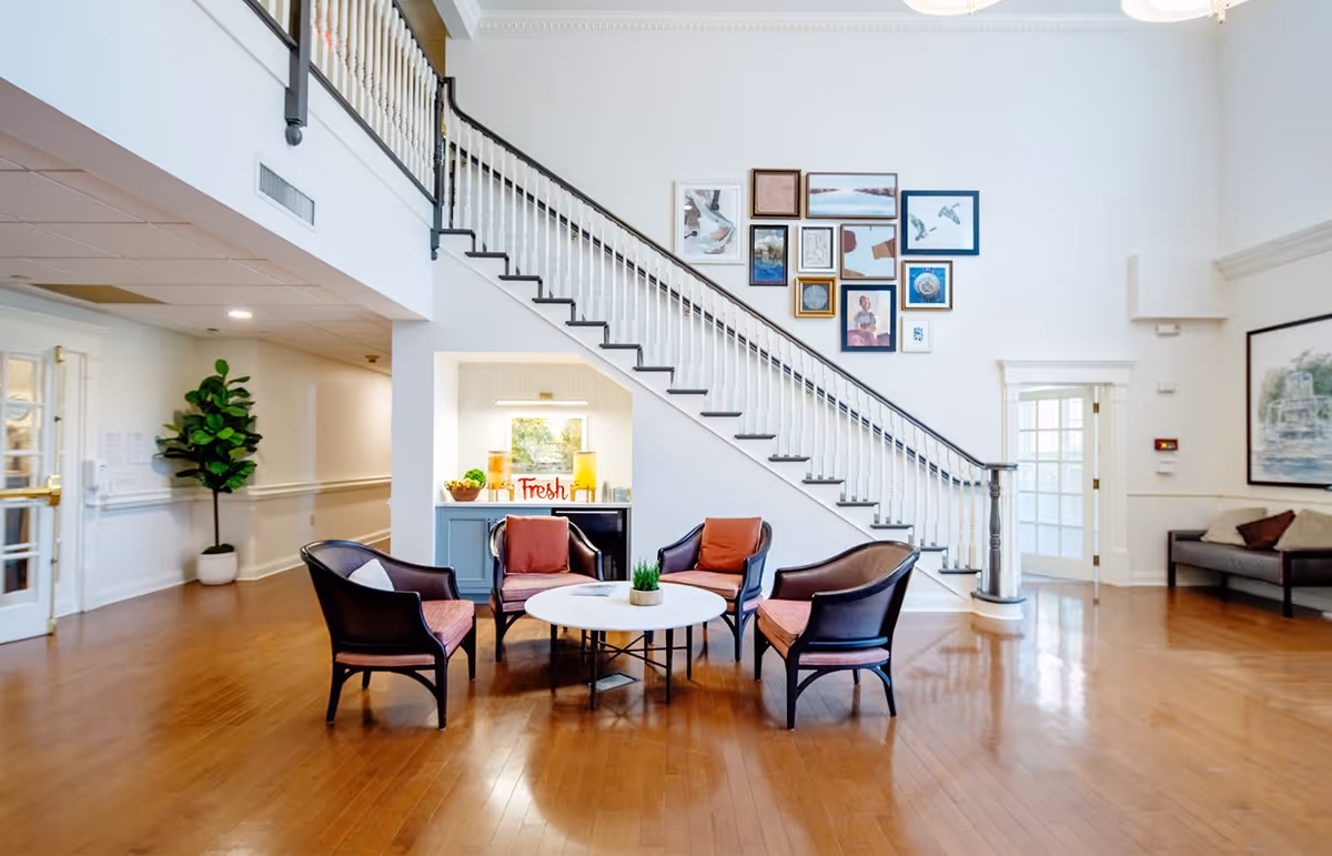 A bright and spacious common area with wooden flooring, featuring a round white table surrounded by four cushioned chairs. A staircase with white railings and dark handrails ascends along the wall decorated with multiple framed artworks. There is a small cabinet with a 'Fresh' sign and decorative items underneath the staircase. A potted plant is placed near a hallway entrance, and a sofa with cushions is visible on the right side of the room.