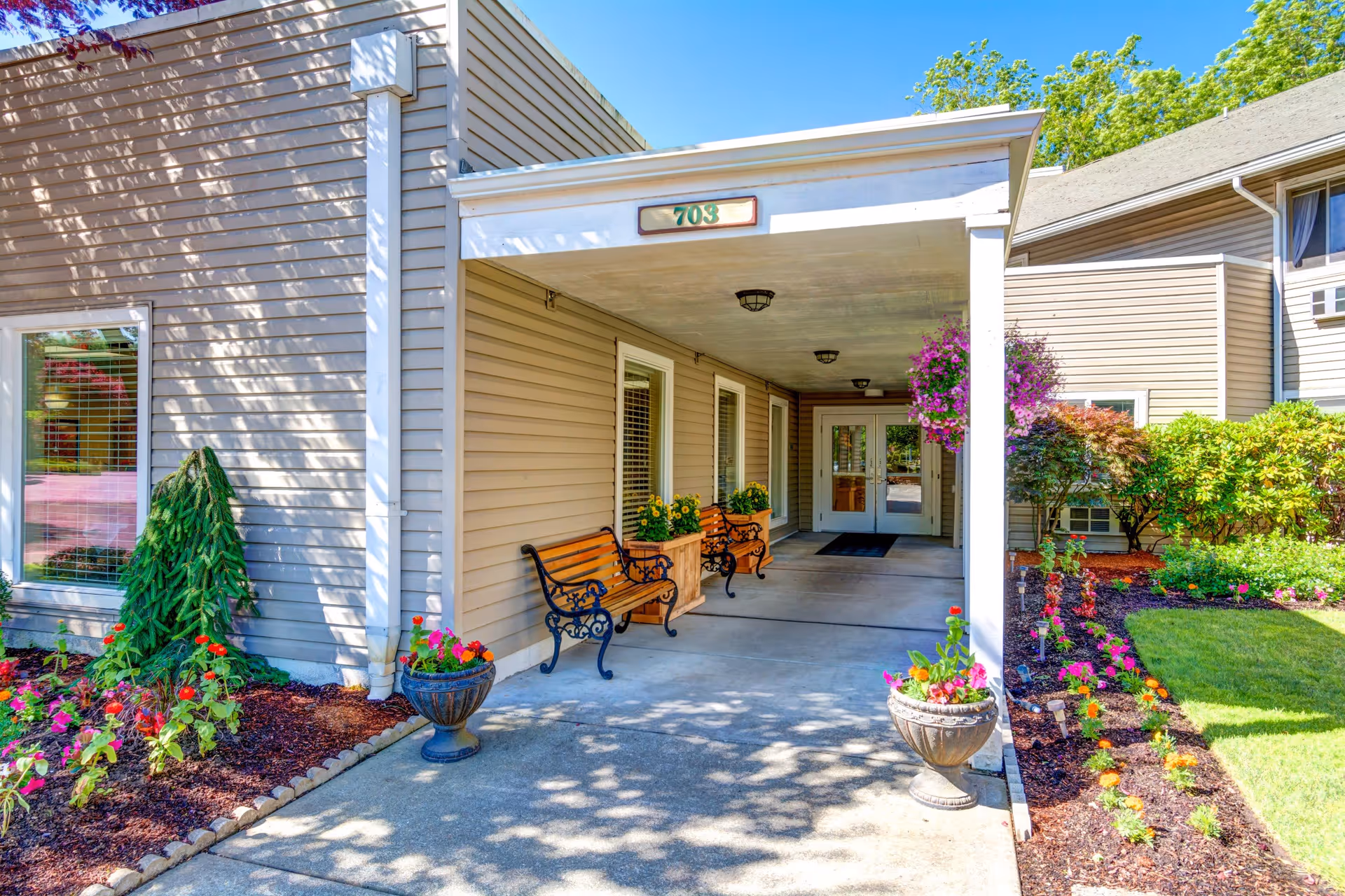 Entrance to a senior living facility with a covered walkway, two wooden benches with black metal armrests, potted flowers, and well-maintained garden beds with colorful flowers and greenery. The building exterior is beige with white trim, and the door has glass panels. The number 703 is displayed above the entrance.