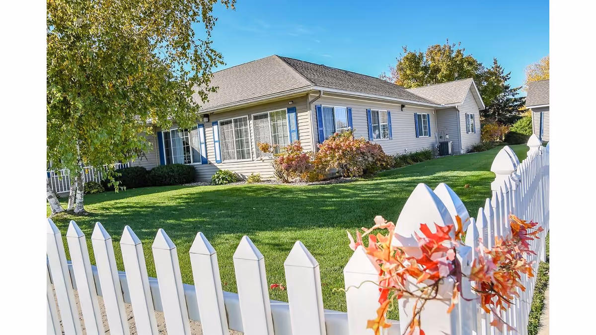 Single-story beige cottage with blue shutters, a manicured lawn and a white picket fence decorated with autumn leaves.