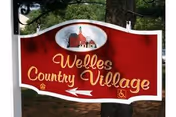 A red and white sign hanging outdoors from a black metal bracket attached to a tree. The sign reads 'Welles Country Village' in yellow cursive letters with a small illustration of a red building with a steeple at the top. There is a white arrow pointing left and a wheelchair accessibility symbol on the bottom right of the sign.