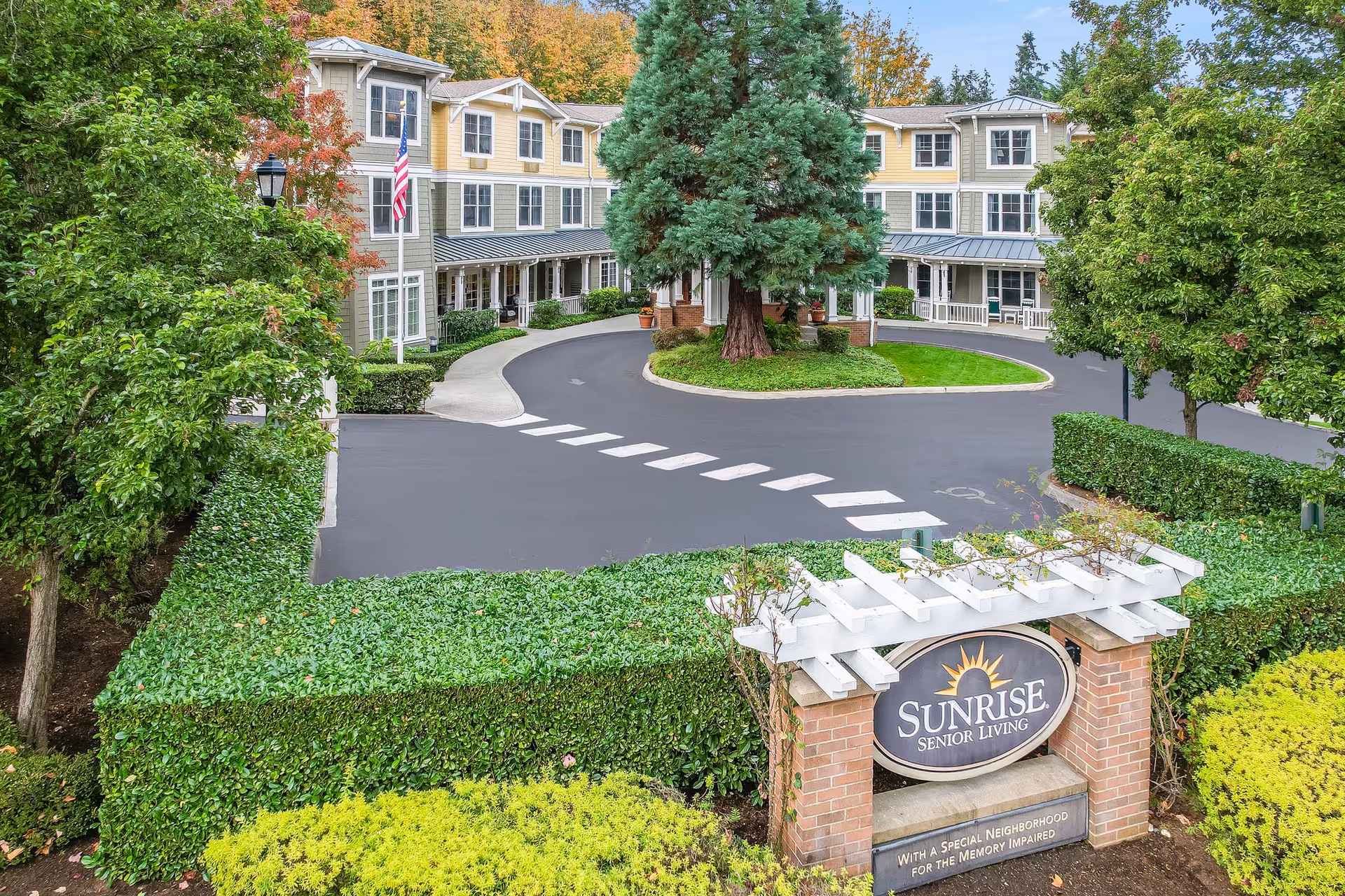Exterior view of Sunrise Senior Living facility in Edmonds, showing a three-story building with yellow and gray siding, surrounded by trees and greenery. There is a circular driveway with a large tree in the center and an American flag on a flagpole near the building. In the foreground, a brick sign with white trellis reads 'Sunrise Senior Living' and 'With a special neighborhood for the memory impaired.'