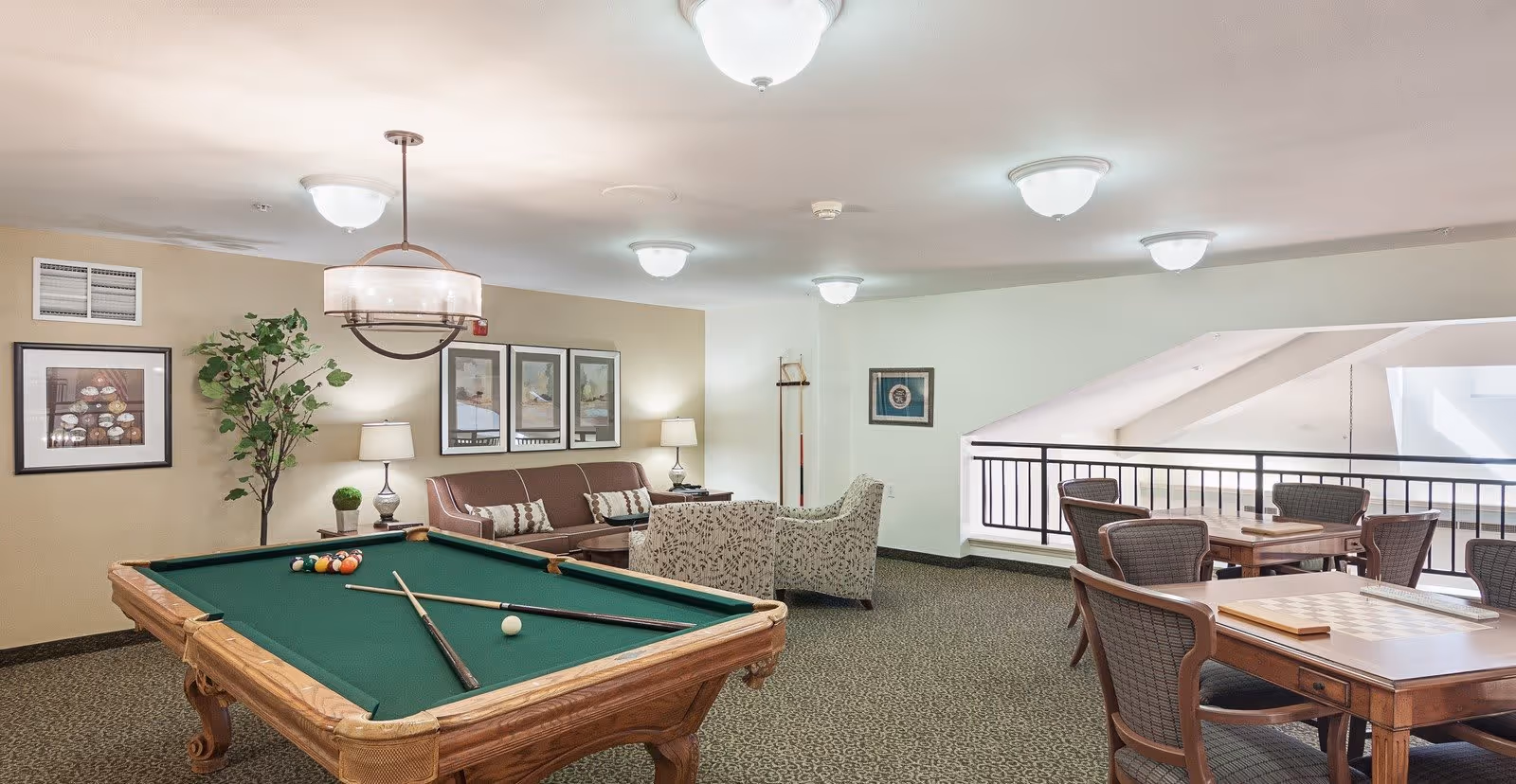 A recreational room with a pool table in the foreground, a brown sofa with patterned cushions, two armchairs, framed artwork on the walls, a potted plant, and several tables with chairs for playing board games. The room has carpeted flooring and ceiling lights.