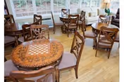 Sunlit dining room with round wooden tables, carved chairs, and a checkerboard tabletop centerpiece.