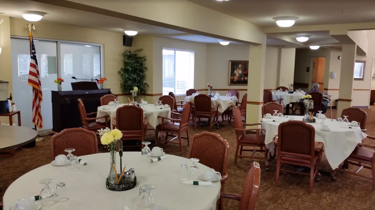 Dining room in a senior living facility with round tables covered with white tablecloths, set with cups, glasses, and napkins. Several chairs are arranged around each table. There is an American flag and a piano in the background, along with a large plant and a framed painting on the wall. The room is well-lit with ceiling lights and has carpeted floors.
