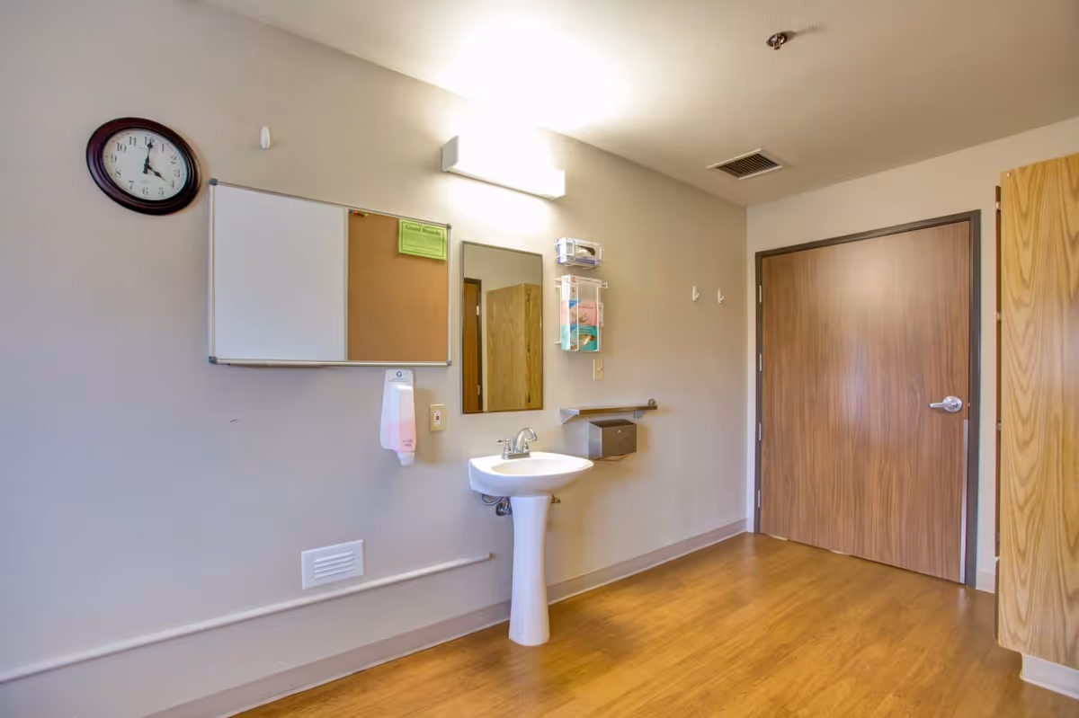 A clean room with a pedestal sink, a wall-mounted mirror, a clock, a whiteboard and corkboard combination, a soap dispenser, and a wooden door. The floor is wood laminate and the walls are painted light beige.