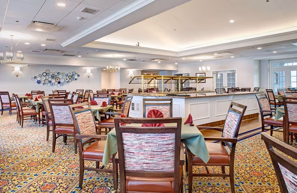 Large senior dining room with multiple set tables and chairs surrounding a central buffet counter under recessed lighting.