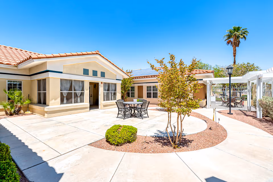 Outdoor courtyard area of a senior living facility with beige buildings featuring red tile roofs, a round metal table with four chairs, small bushes, a tree, and a white pergola with additional seating under a clear blue sky.