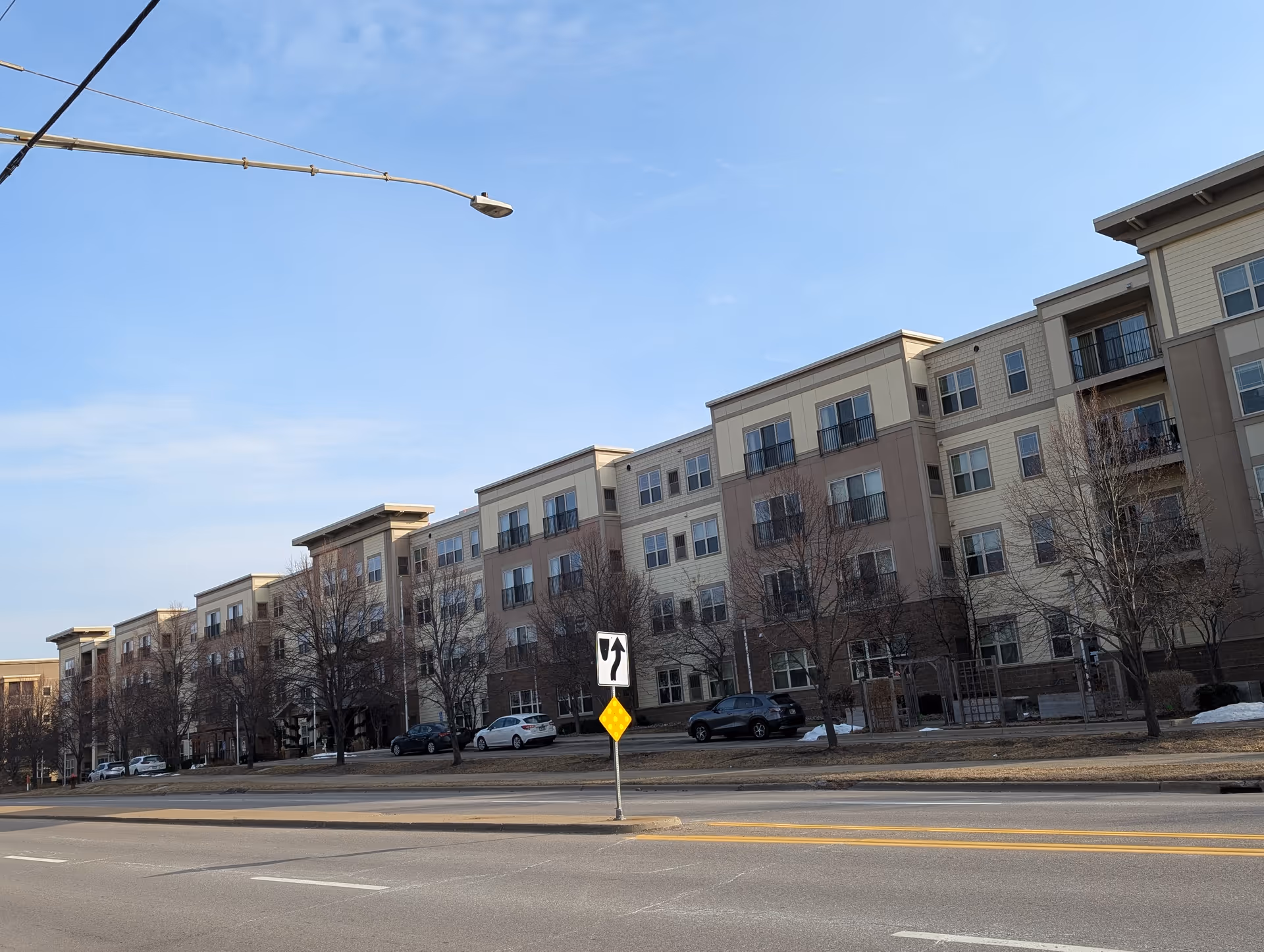Exterior view of a multi-story senior living facility building with several windows and balconies, situated along a street with a traffic sign and parked cars in front. Leafless trees are visible along the sidewalk under a clear blue sky.