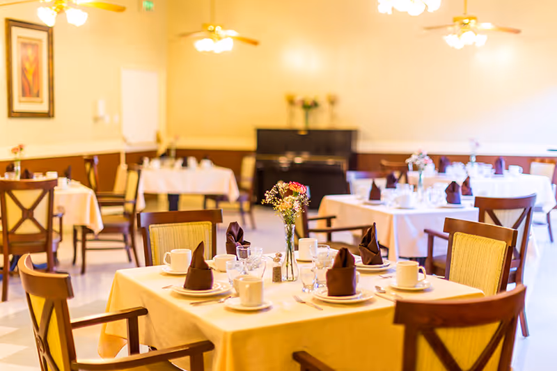 A dining room set up with tables covered in white tablecloths, each table set with cups, plates, glasses, and brown folded napkins. There are wooden chairs with cushioned seats around the tables. The room is warmly lit with ceiling fans and lights, and a piano is visible in the background.